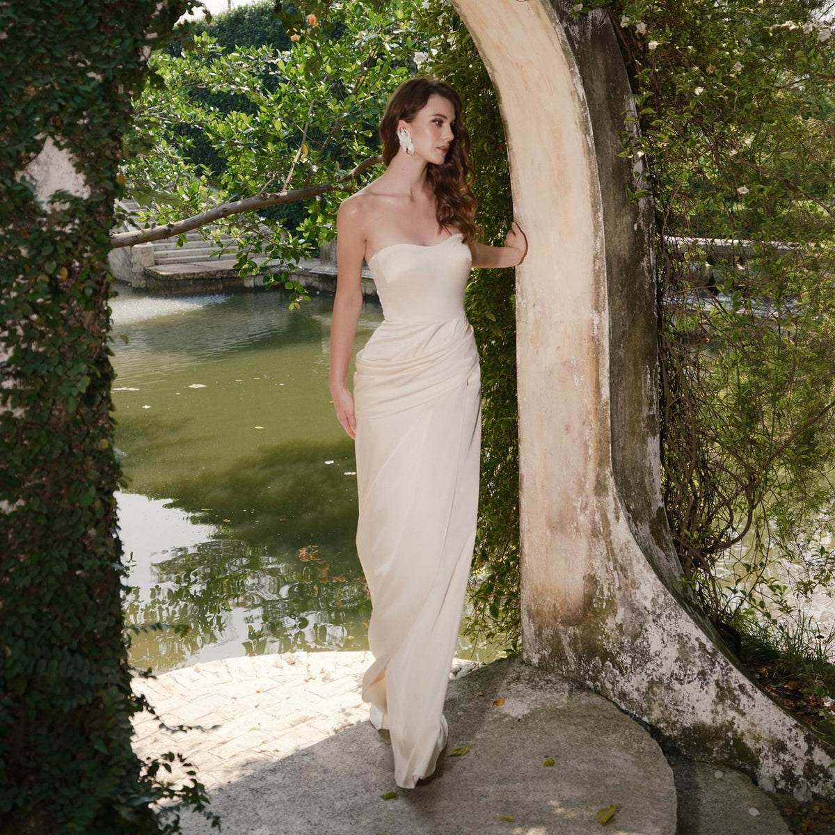 May include: A strapless, floor-length ivory gown with a draped bodice and a flowing skirt. The dress is worn outdoors near a body of water, framed by greenery and a stone archway. The overall aesthetic is elegant and sophisticated.