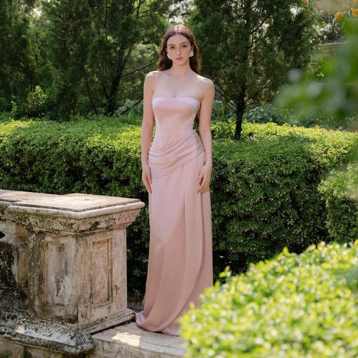 May include: A strapless, floor-length blush pink gown. The dress features a draped bodice and a flowing skirt with a subtle slit. The image is set outdoors with a backdrop of lush greenery and a stone structure.