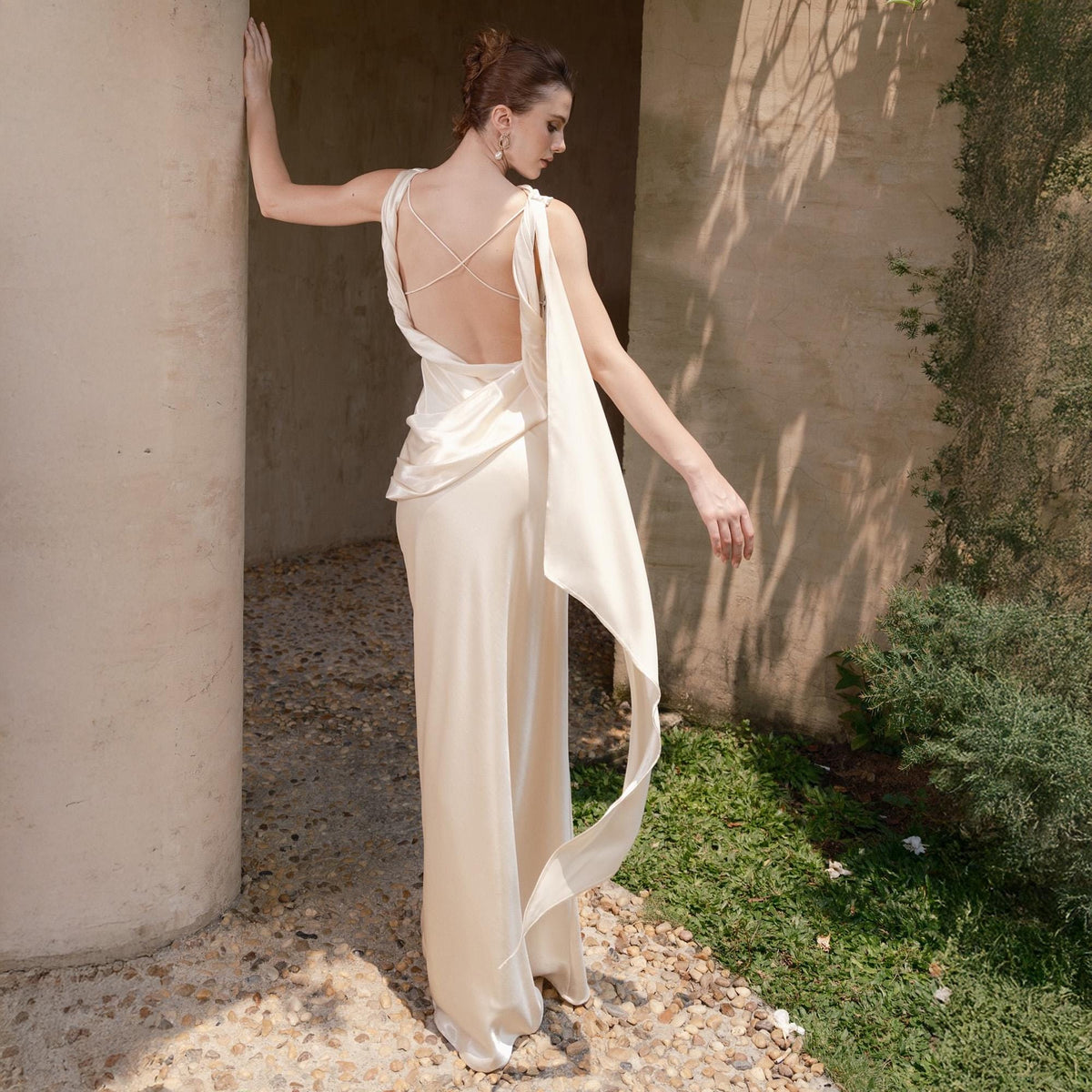 May include: Ivory-colored formal wear ensemble. The top features a crisscross back design and a draped fabric element that flows down the back. The outfit includes wide-leg pants. The model is standing near a stone column and greenery.