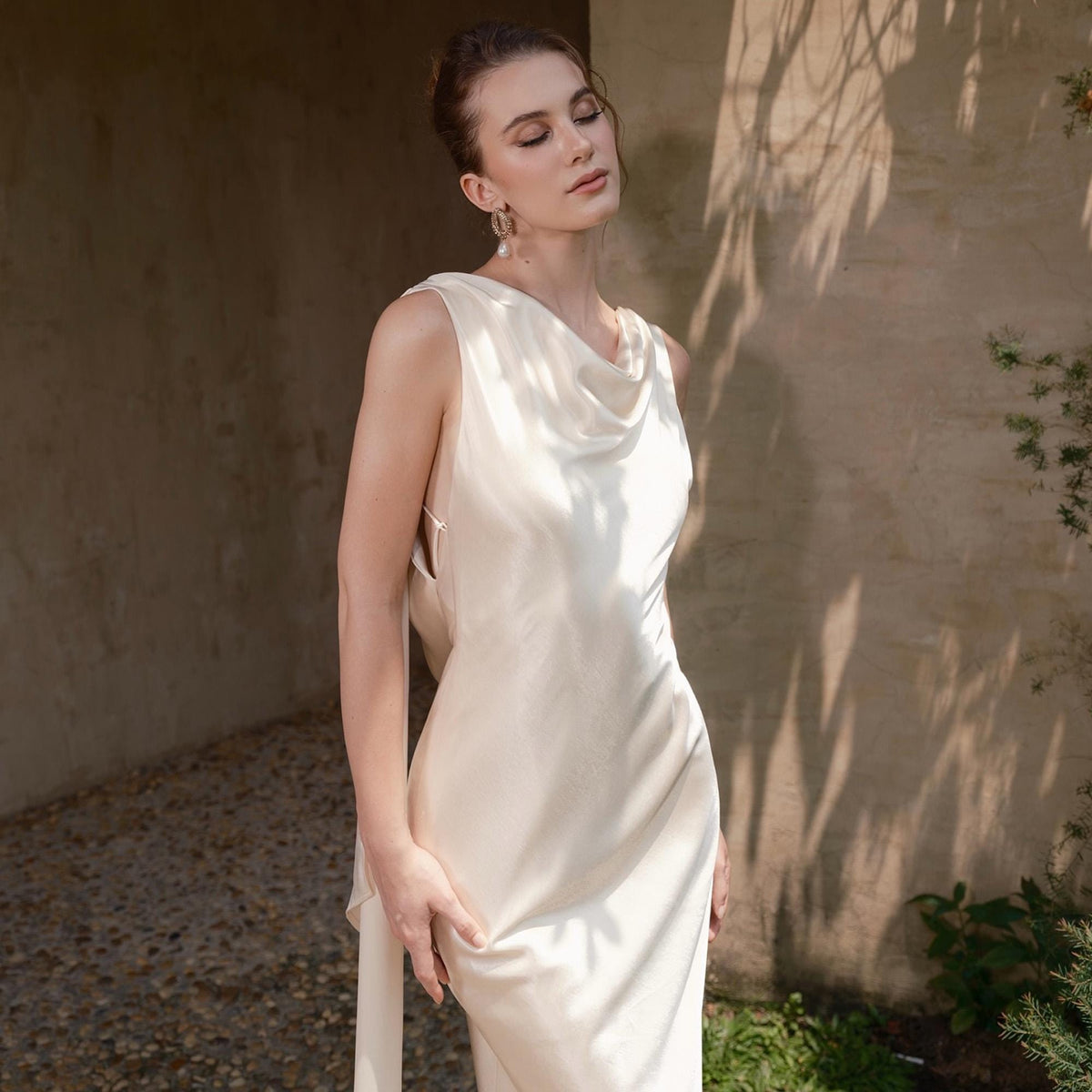 May include: A sleeveless, ivory-colored satin dress with a cowl neckline. The dress has a flowing silhouette and a long, elegant train. The model is wearing gold earrings and has her eyes closed.