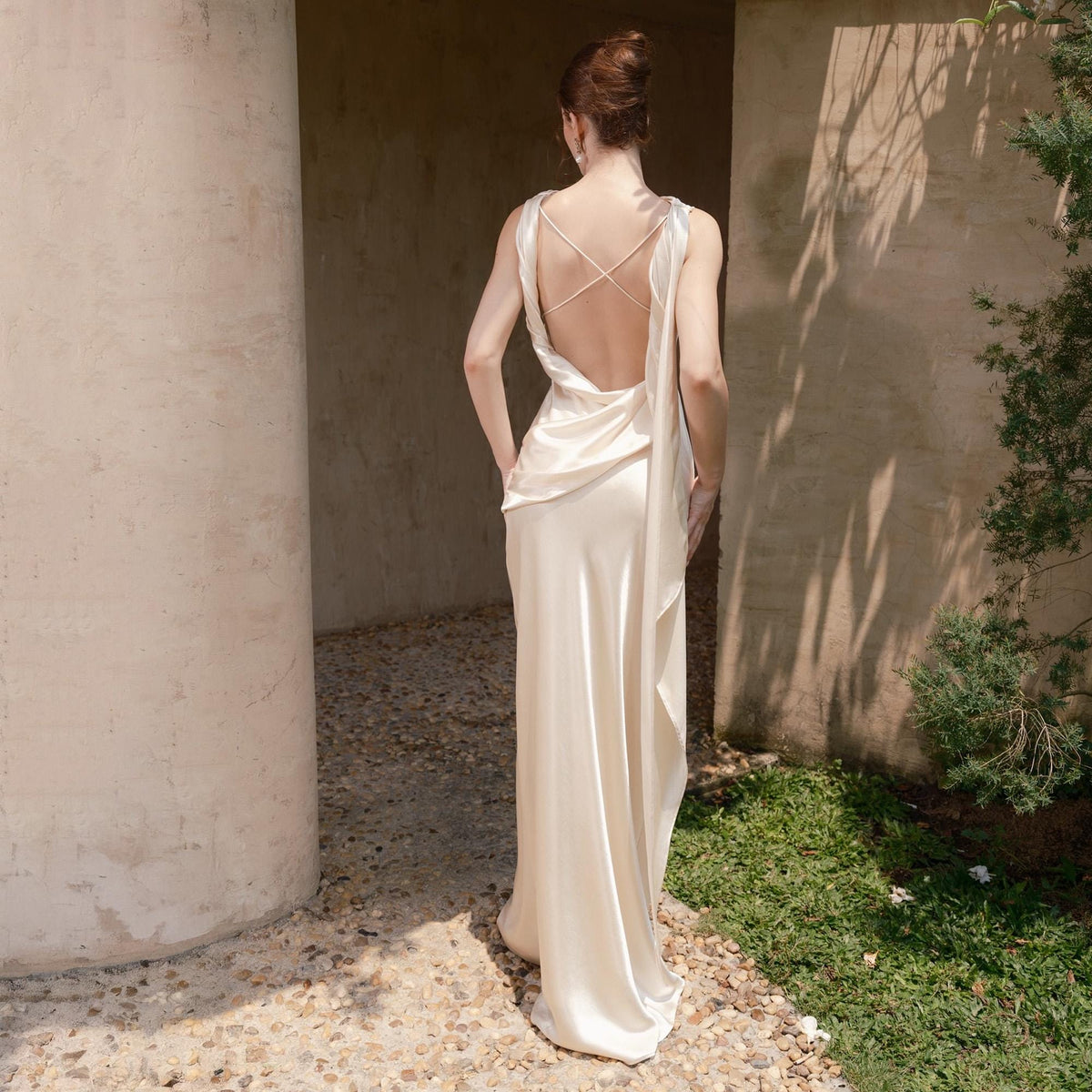 May include: Ivory-colored, floor-length gown with a draped back and a crisscross strap design. The dress has a flowing, silky texture and a subtle sheen. The model is standing outdoors near a stone wall and greenery.
