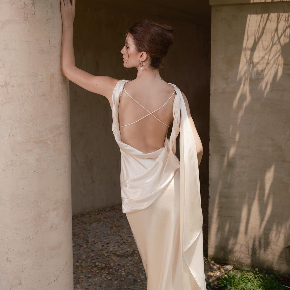 May include: A cream-colored, backless gown with spaghetti straps that crisscross the back. The dress has a draped design and a flowing skirt. The model is leaning against a column in an outdoor setting. The dress is made of a silky material.