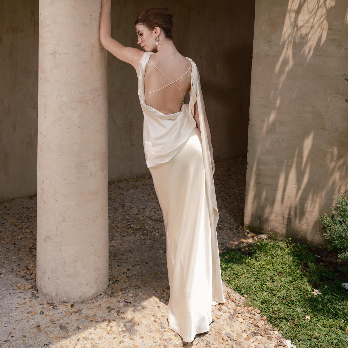 May include: A cream-colored, floor-length gown with a draped bodice and open back. The dress features delicate spaghetti straps that cross in the back and a flowing skirt. The gown is made of a silky material and has a subtle sheen.