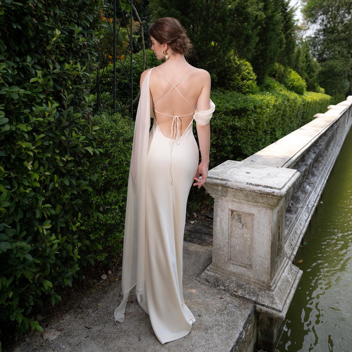May include: A cream-colored, backless formal gown with a flowing train and delicate straps. The dress features a draped sleeve and is worn outdoors near a stone wall and greenery.