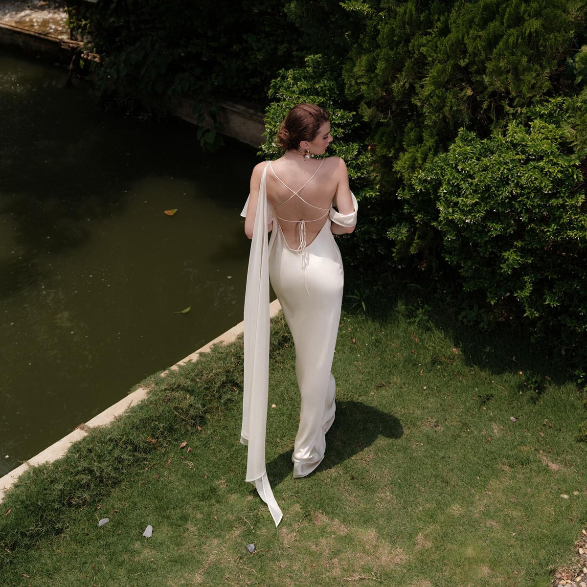 May include: A white, floor-length gown with a low back and a flowing train. The dress features delicate straps that cross the back and drape over the shoulders. The gown is worn outdoors near a body of water and green foliage.