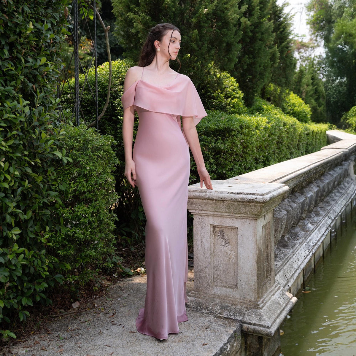 May include: A light pink, floor-length gown with a draped, off-the-shoulder neckline. The dress features a flowing skirt and a delicate, layered bodice. The model is standing outdoors near a stone wall and greenery.
