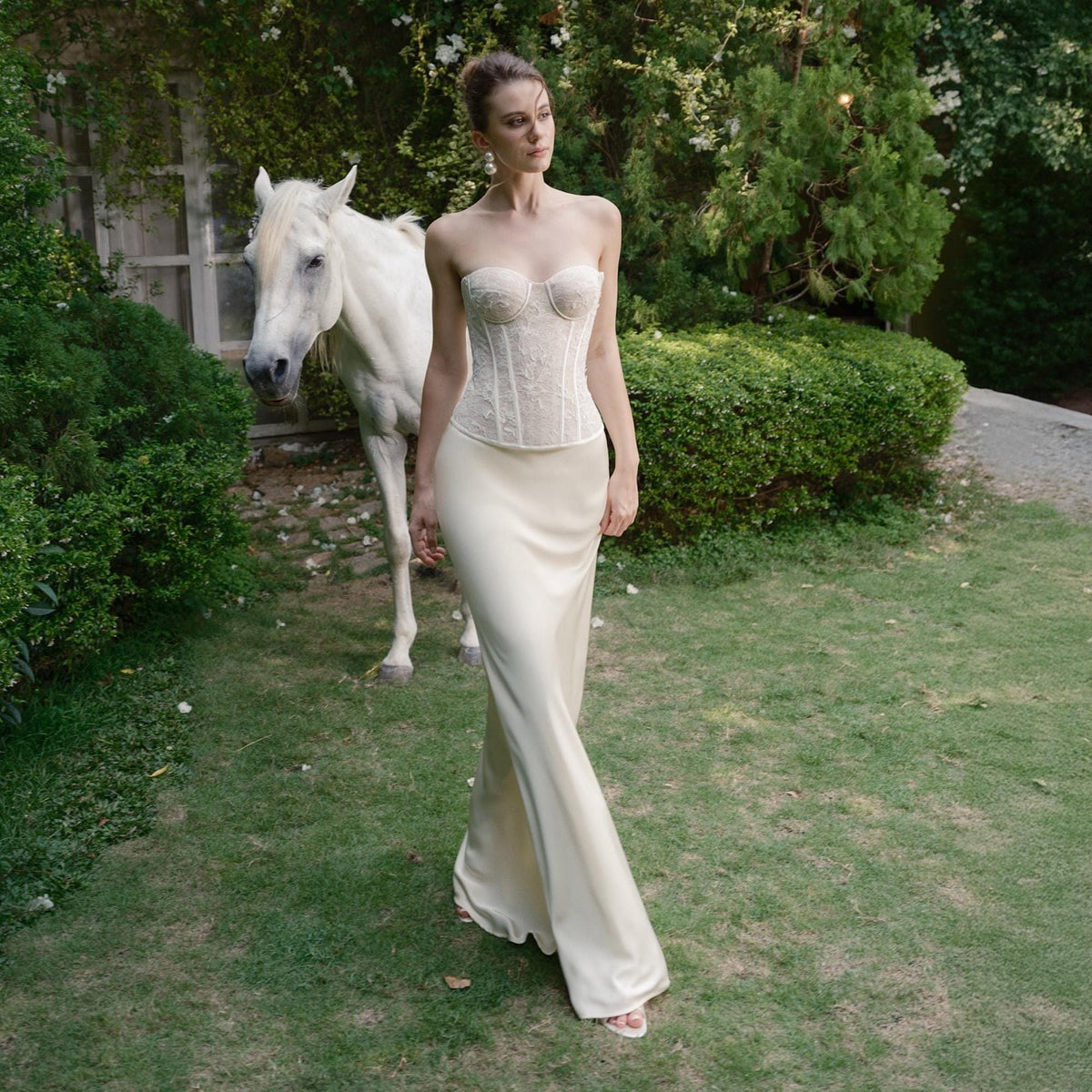 May include: A strapless ivory corset top with delicate lace detailing paired with a matching floor-length satin skirt. A white horse stands in the background, with lush greenery surrounding the scene. The overall aesthetic is elegant and romantic.