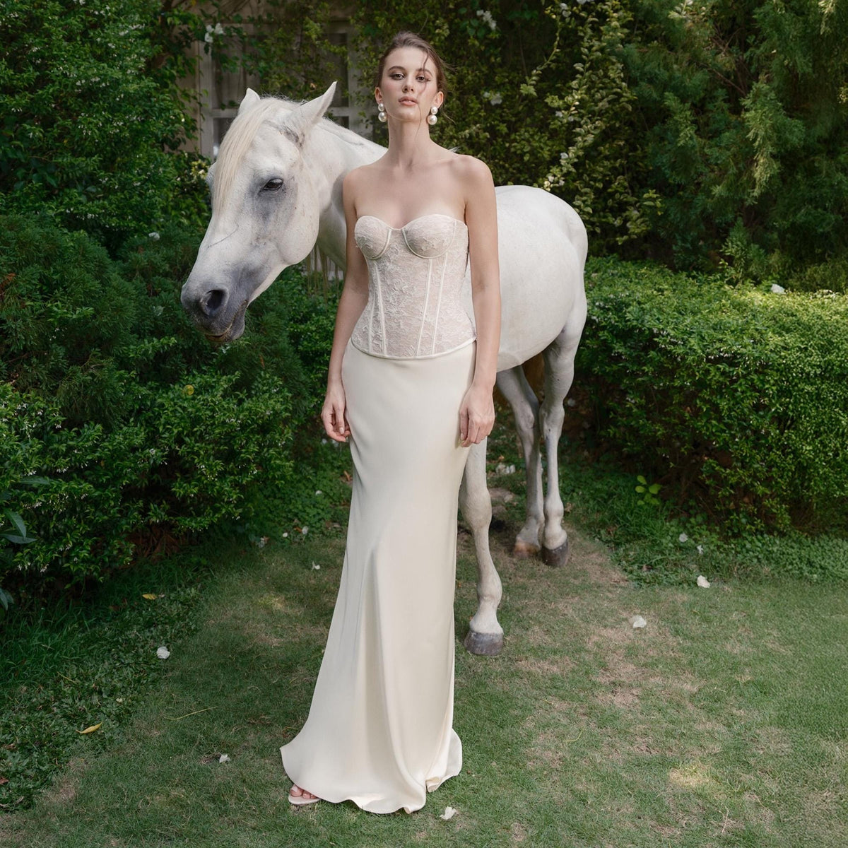 May include: A strapless ivory corset top with lace detailing paired with a matching floor-length skirt. A white horse stands behind the person, set against a backdrop of green foliage and grass.