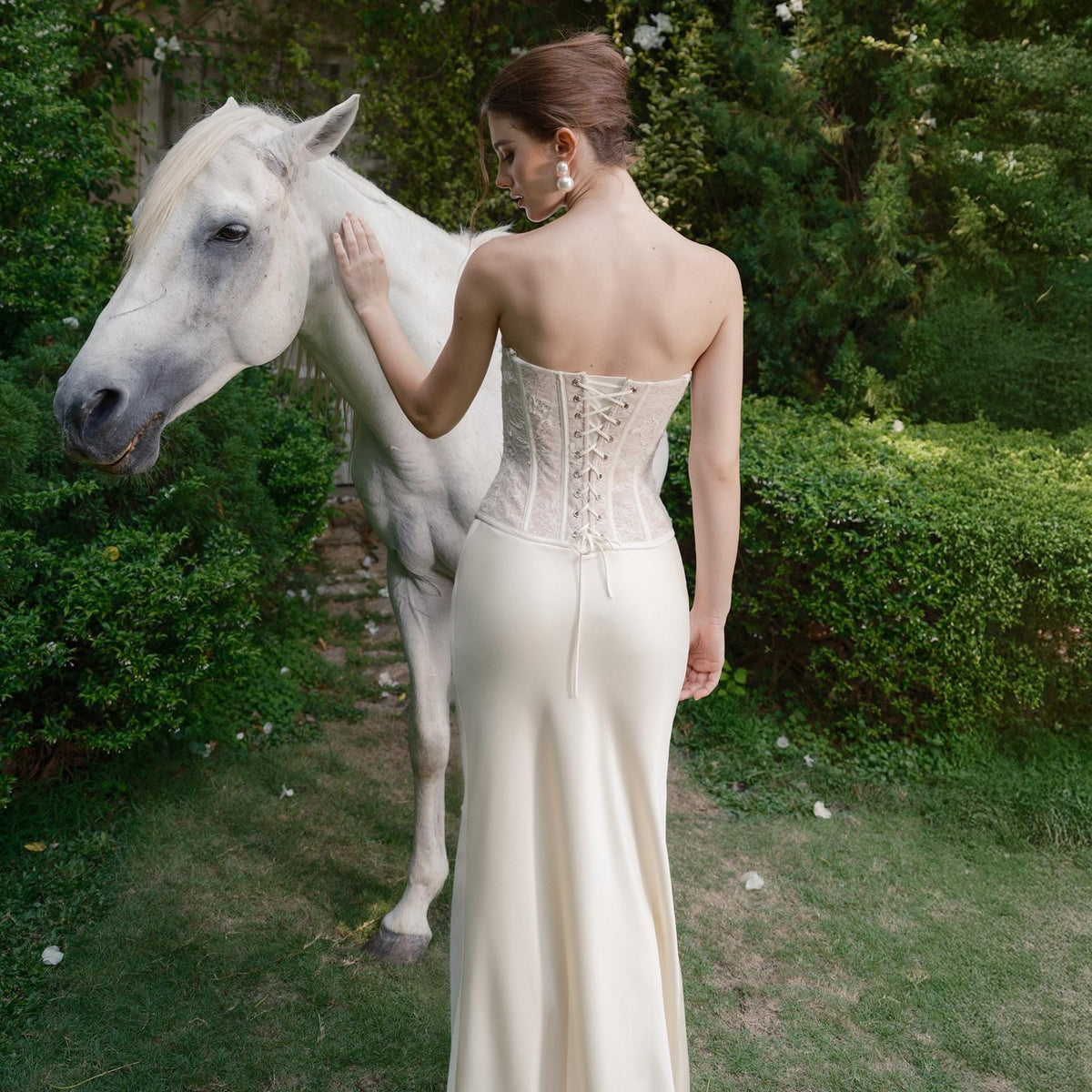 May include: A strapless ivory corset with lace detailing and a matching floor-length skirt. A white horse stands nearby in a garden setting. The corset features a lace-up back. The scene is set outdoors with green foliage.