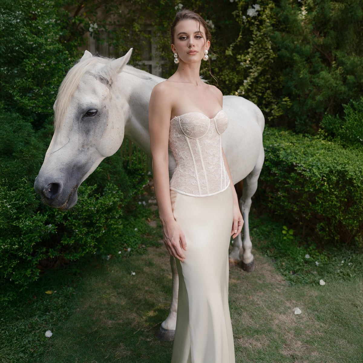 May include: A strapless ivory corset top with delicate lace detailing and a matching floor-length skirt. A white horse stands behind the model in a garden setting. The model is wearing pearl earrings.