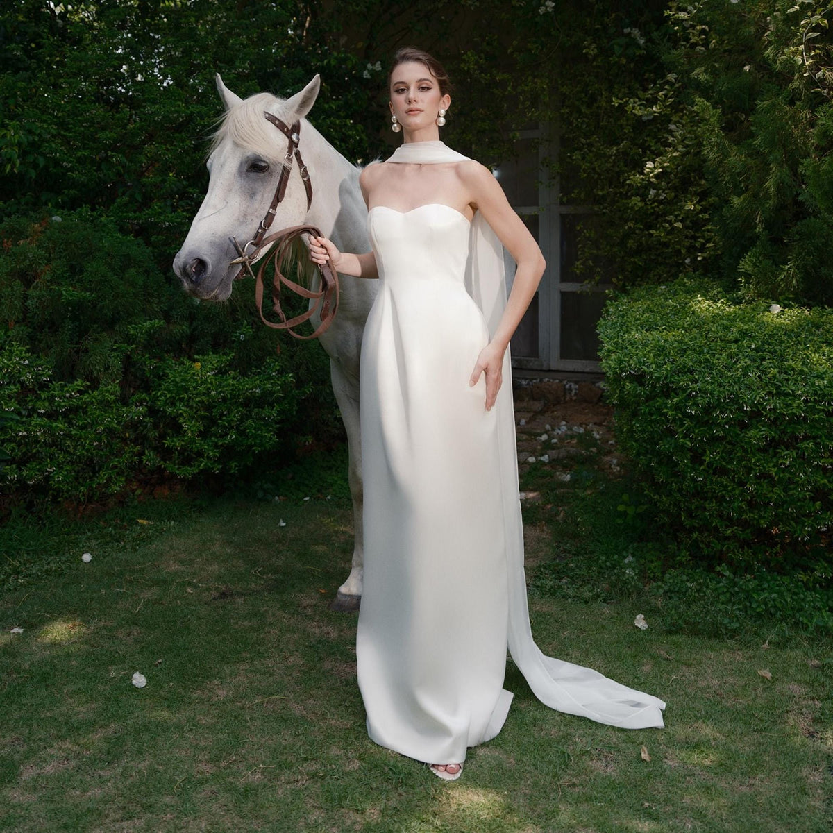 May include: A woman in a white strapless gown with a flowing cape stands next to a white horse. The dress is floor-length and has a fitted bodice. The horse has a brown bridle and reins. The scene is set outdoors with green foliage in the background.