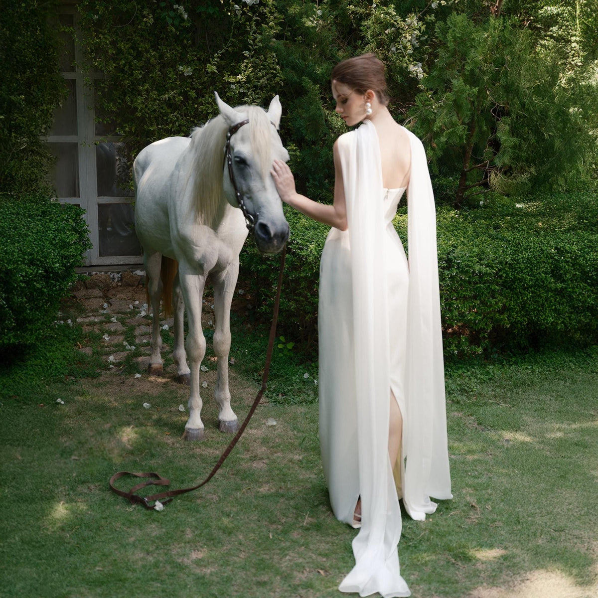 May include: A white, floor-length gown with a cape detail. A woman stands next to a white horse in a garden setting. The horse has a bridle and a brown leather lead. The woman is wearing a white dress with a high slit and a cape that drapes over one shoulder.