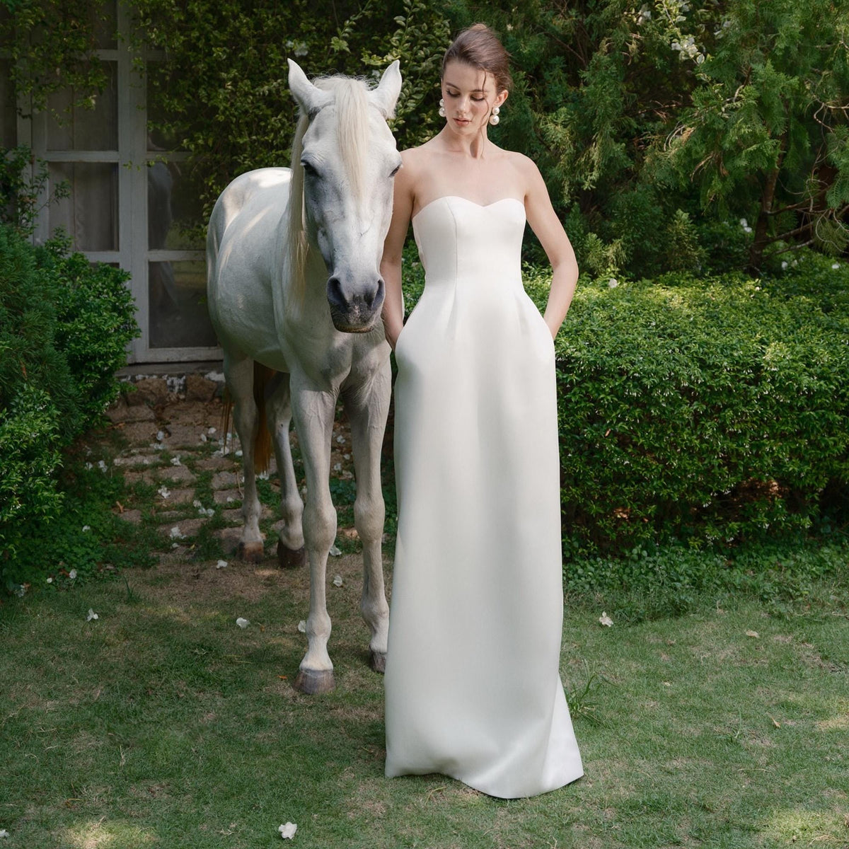 May include: A strapless, floor-length white gown with pockets. A white horse stands beside the person wearing the dress in a garden setting. The dress has a simple, elegant design.