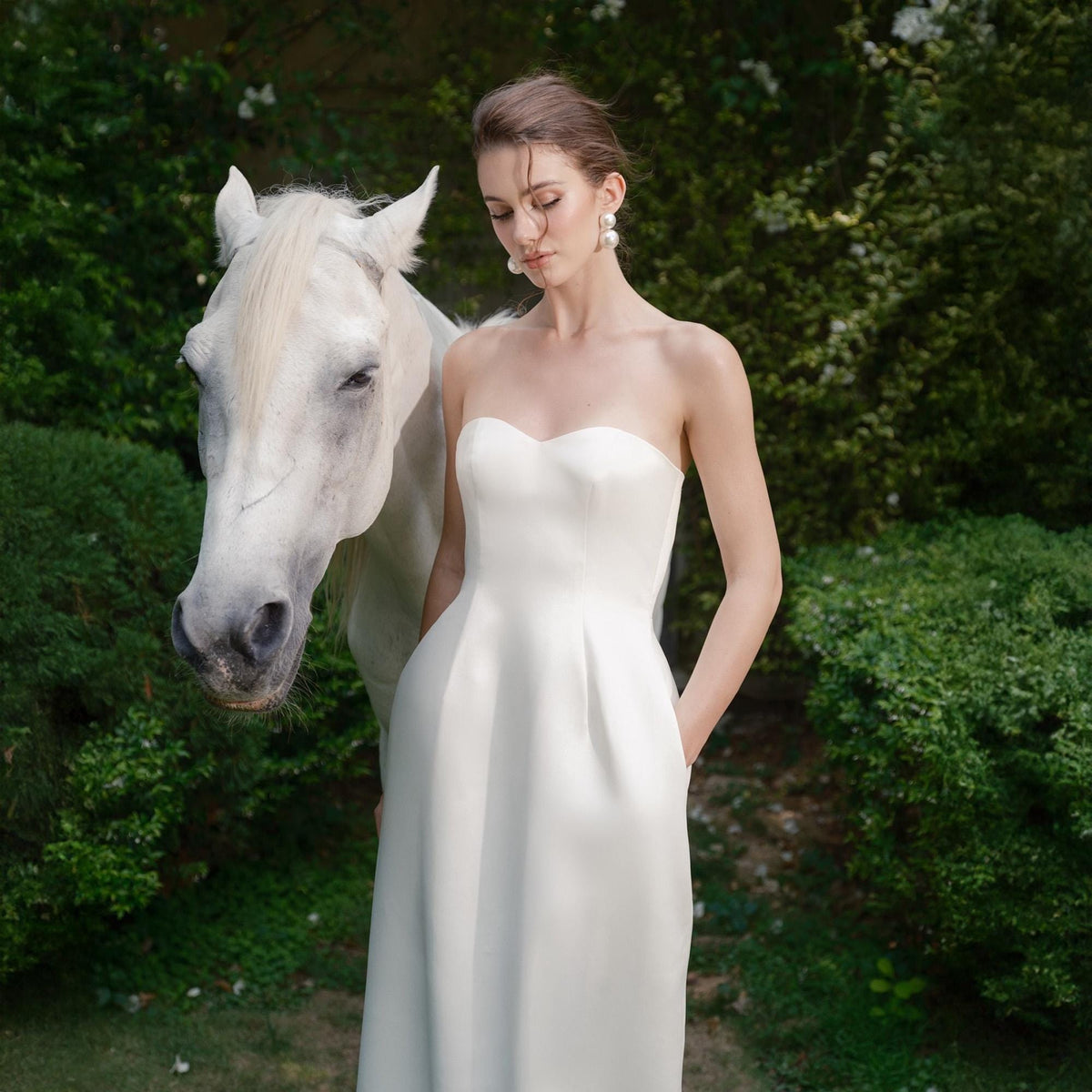 May include: A strapless white dress with a sweetheart neckline. The dress is worn by a person standing next to a white horse. The background features green foliage. The person is wearing large pearl earrings.