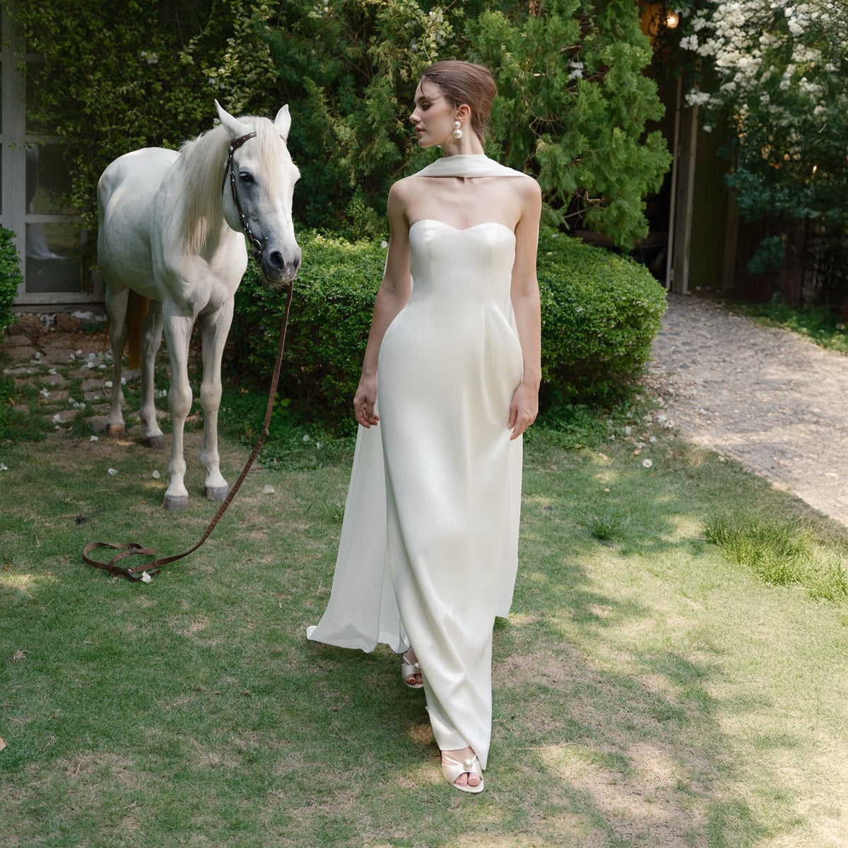 May include: A strapless, ivory-colored gown with a flowing skirt. A white horse stands nearby on a grassy lawn. The dress features a sweetheart neckline and a matching scarf. The scene is set outdoors with greenery in the background.