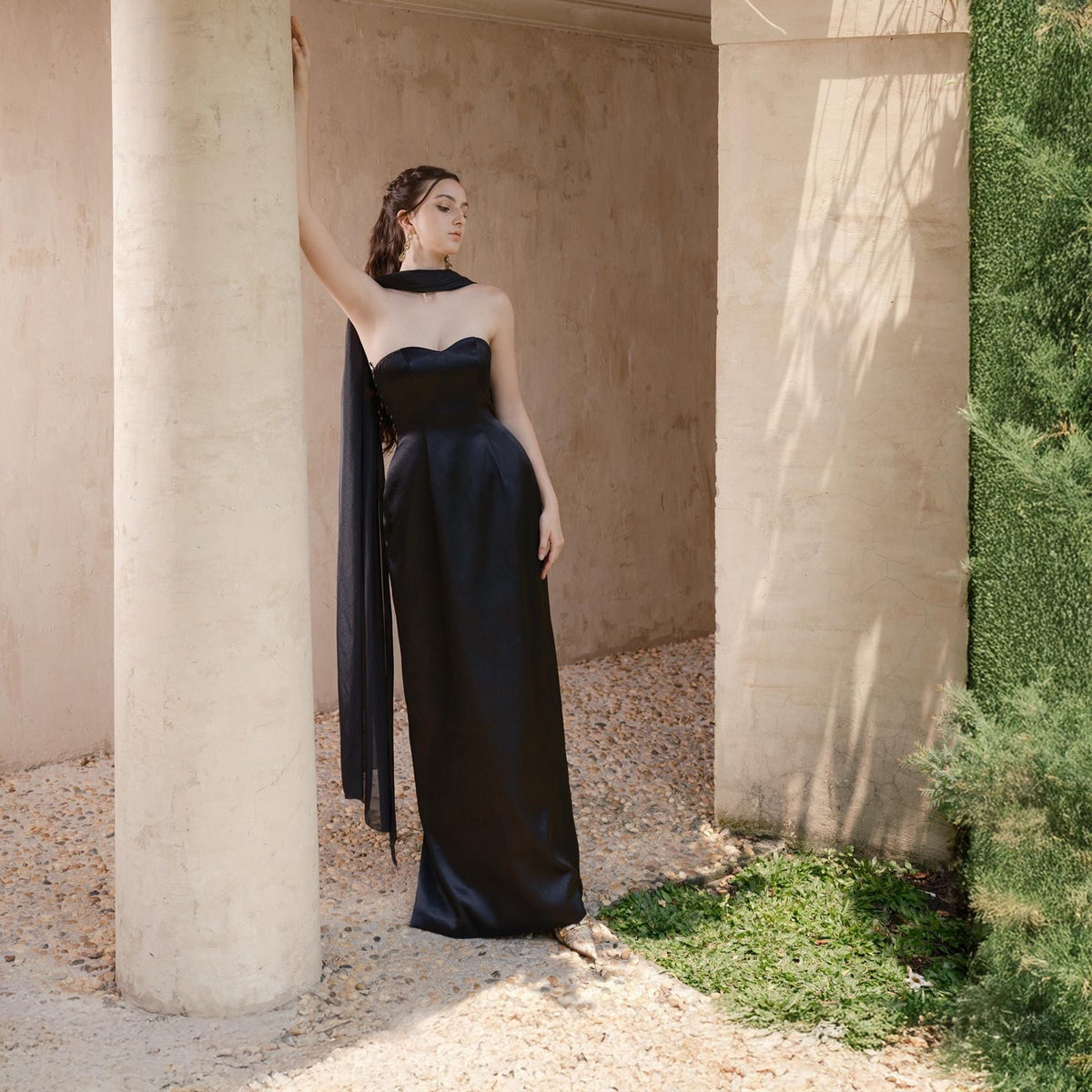May include: A strapless, floor-length black gown with a flowing scarf detail. The dress has a fitted bodice and a sleek, elegant silhouette. The background features architectural elements and greenery.