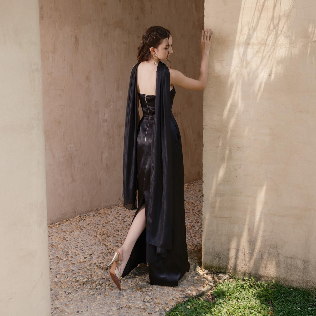 May include: A black evening gown with a high slit and attached cape sleeves. The dress has a fitted bodice and a long, flowing skirt. The model is wearing gold-colored heels.