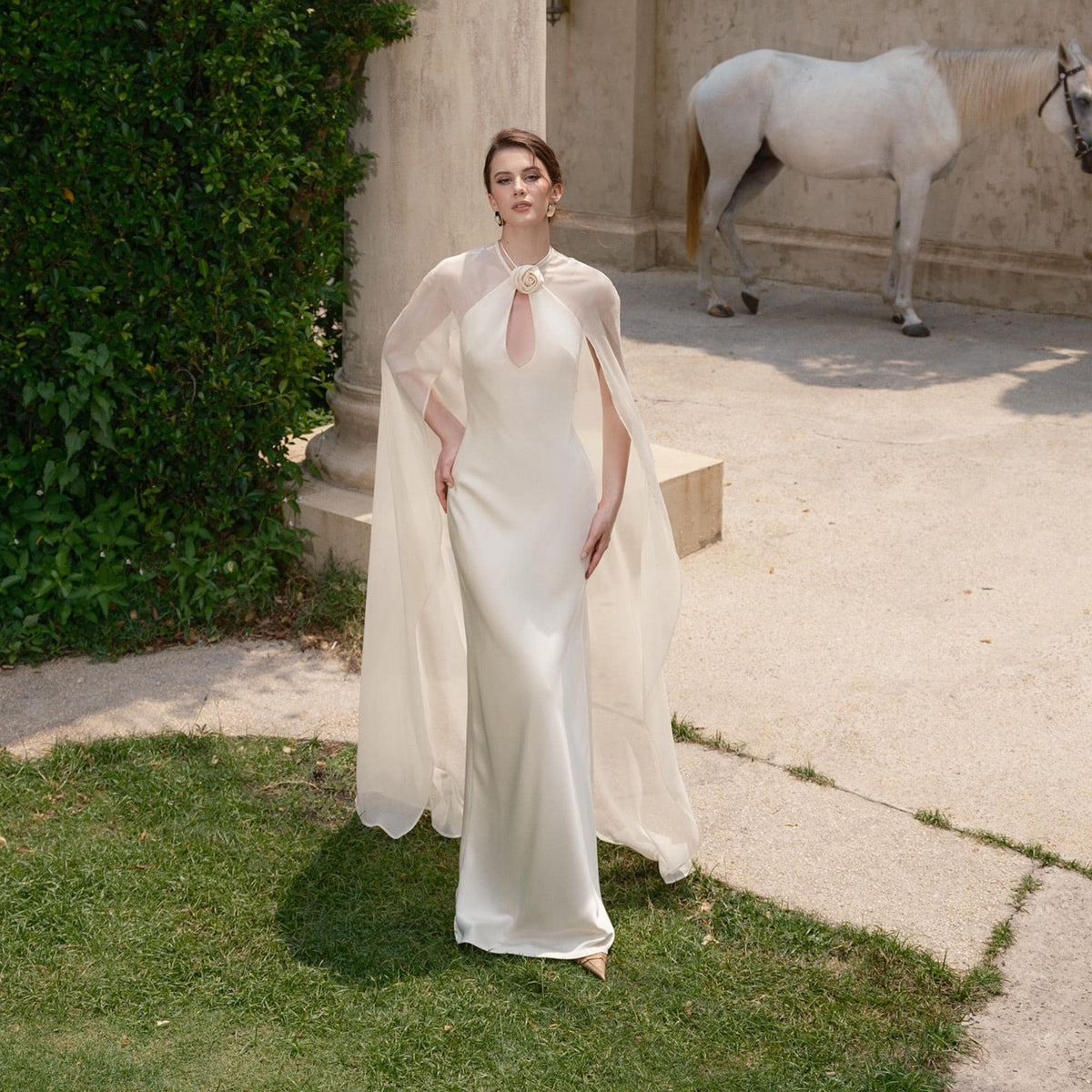 May include: A floor-length ivory gown with a keyhole neckline and a sheer cape. The cape is attached at the shoulders and drapes down the arms. A white flower adorns the neckline. A white horse stands in the background.