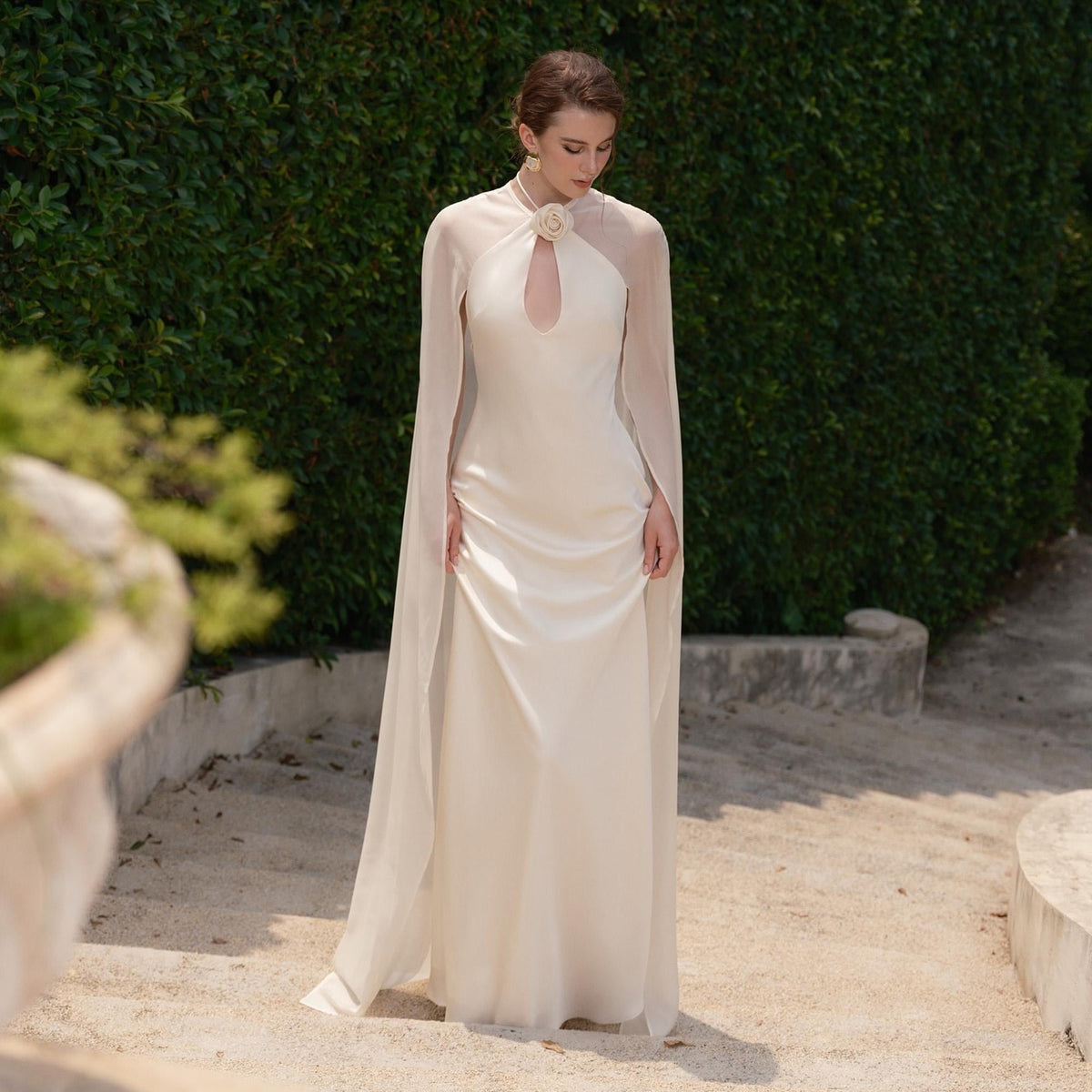 May include: An ivory-colored gown with a keyhole neckline and a sheer cape. The dress features a high neckline with a floral embellishment. The model is standing on a set of stone steps in front of a green hedge.