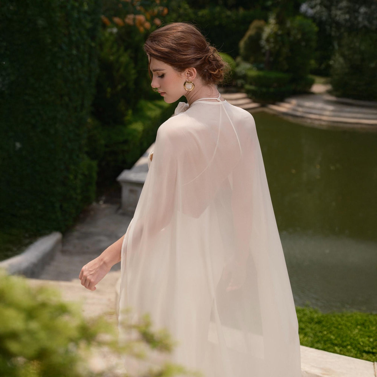 May include: A sheer, ivory-colored cape with a high neckline. The cape is flowing and lightweight, suitable for formal occasions. The background features a garden setting with a pond and greenery.
