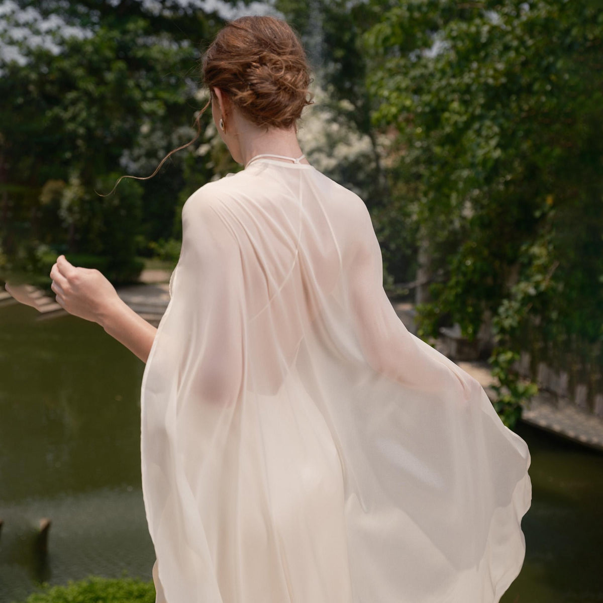 May include: A sheer, cream-colored cape with a high neckline. The cape drapes over the shoulders and arms, creating a flowing silhouette. The background features a blurred view of a body of water and lush greenery.