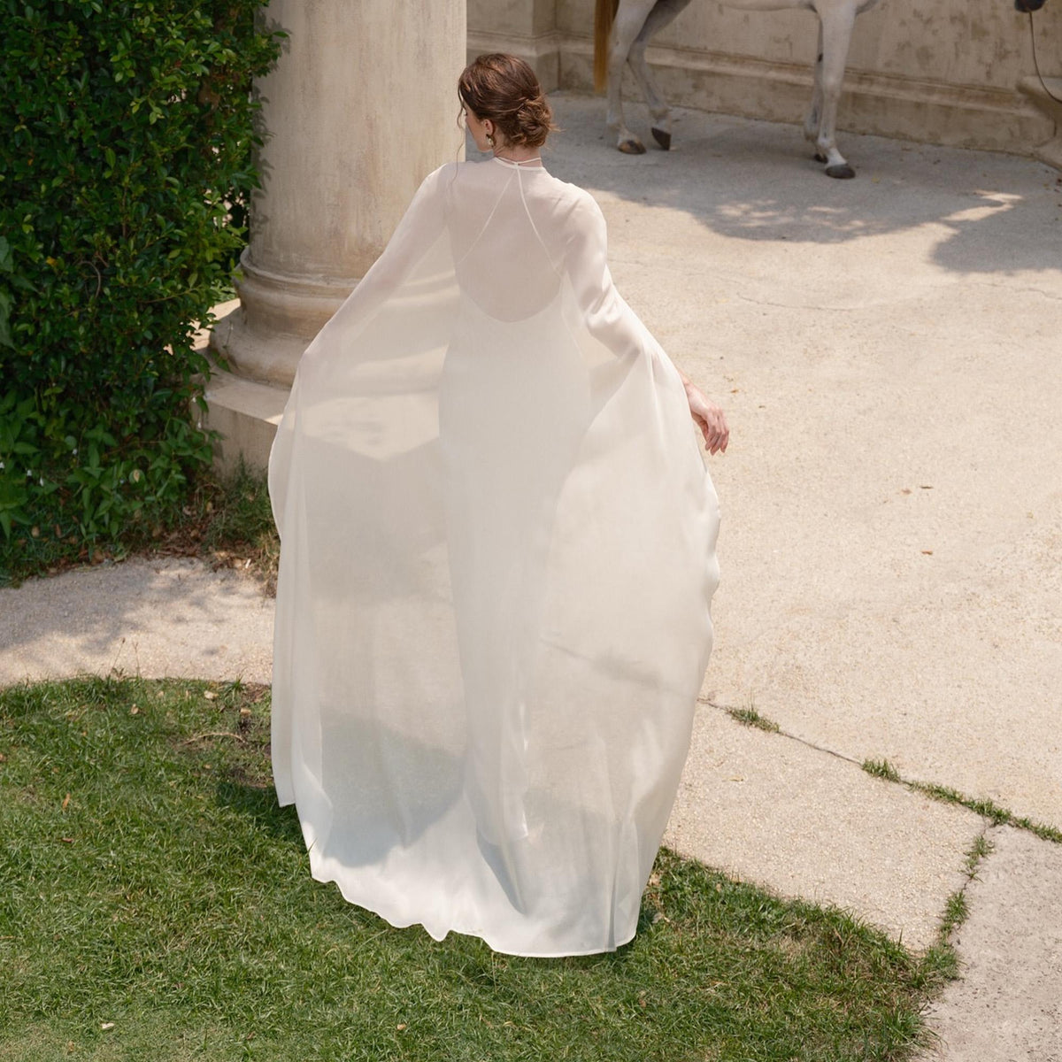 May include: A flowing, white bridal gown with a sheer cape, captured from behind. The dress has a high neckline and a long, trailing cape. The scene is outdoors, with green grass and a stone walkway.