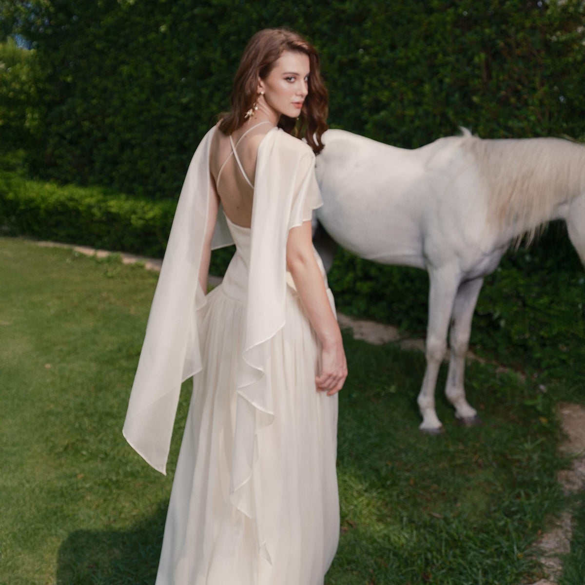 May include: A woman in an ivory-colored gown with flowing cape sleeves stands in a grassy area next to a white horse. The dress has a low back and a flowing skirt. The background features green foliage.