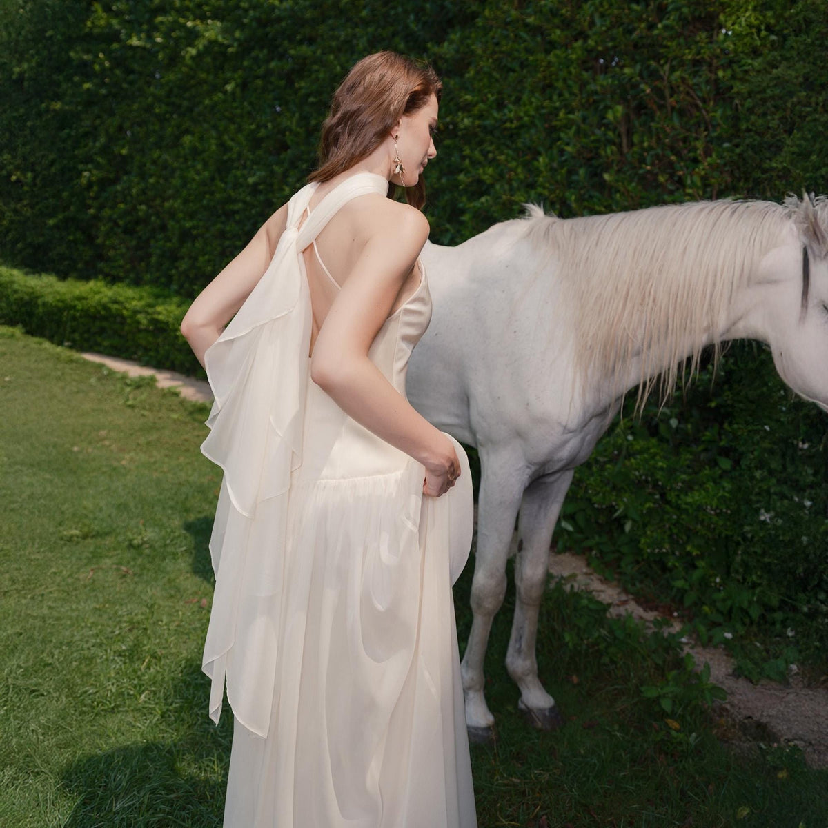 May include: A cream-colored formal gown with a halter neckline and flowing skirt. The dress features a layered, sheer back detail. A white horse stands nearby in a grassy outdoor setting.