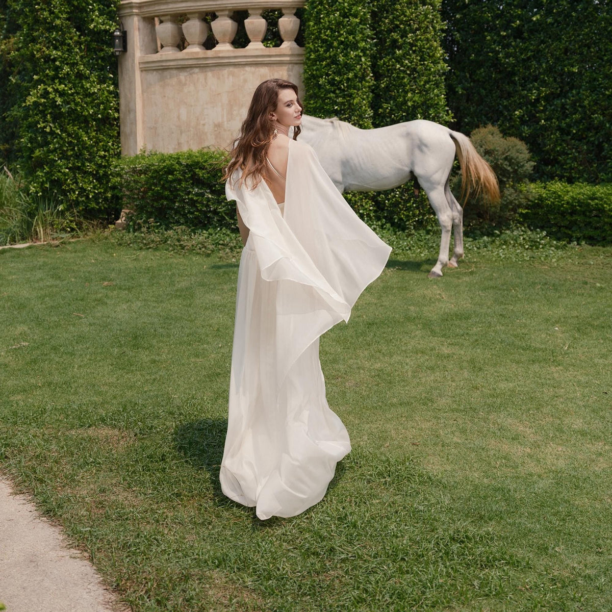 May include: A woman in a flowing, ivory-colored gown with a cape stands on a grassy lawn. A white horse is in the background. The dress has a low back and a long, elegant train. The scene is outdoors with greenery and a stone structure.