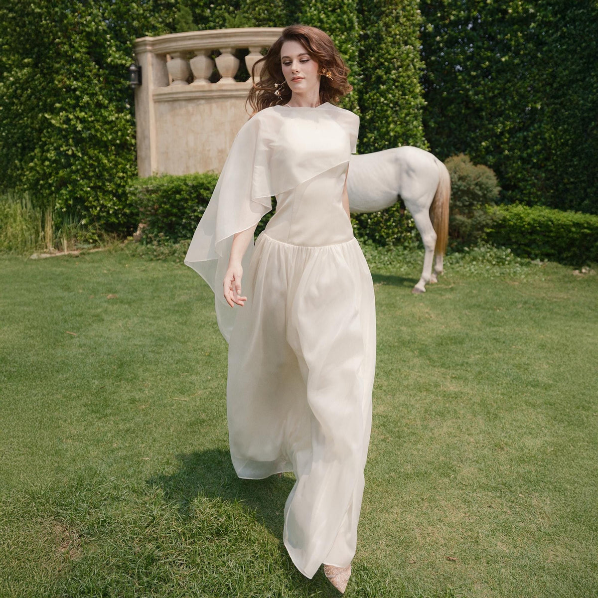 May include: A cream-colored, flowing gown with a cape-like overlay. The dress features a fitted bodice and a full, floor-length skirt. The model is walking on green grass in an outdoor setting, with a white horse statue in the background.
