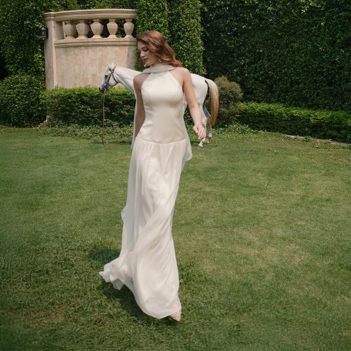 May include: A woman in a cream-colored halter dress walks on a green lawn, with a white horse in the background. The dress has a flowing skirt and a scarf-like detail around the neck. The scene is set in a garden.