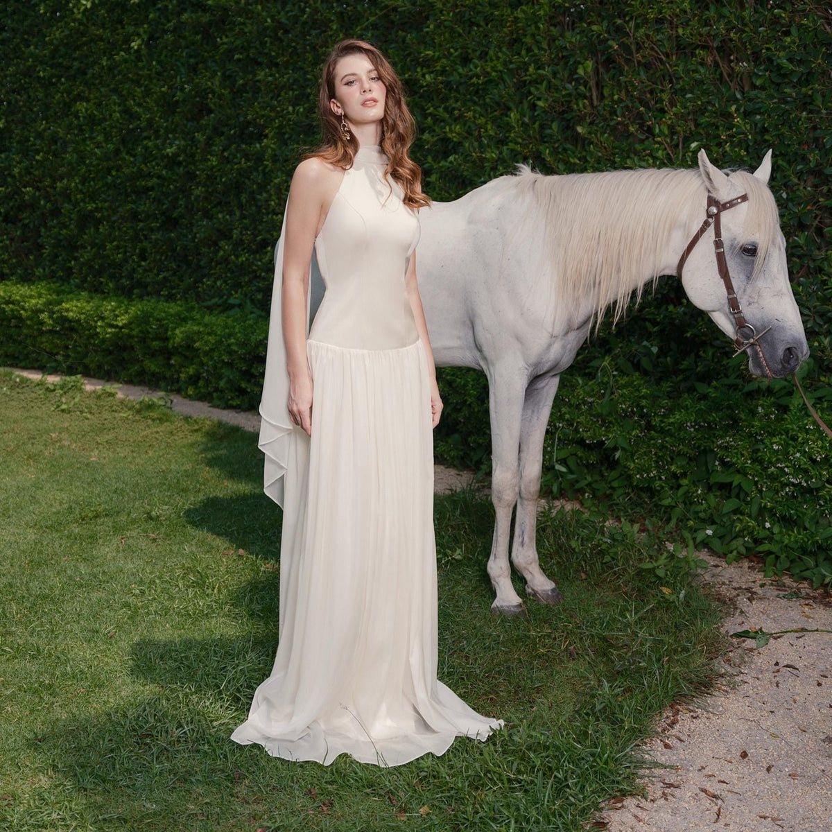 May include: A woman in a cream-colored halter dress with a flowing skirt stands next to a white horse. The dress has a high neckline and a sheer overlay. The scene is set outdoors with green grass and foliage in the background.