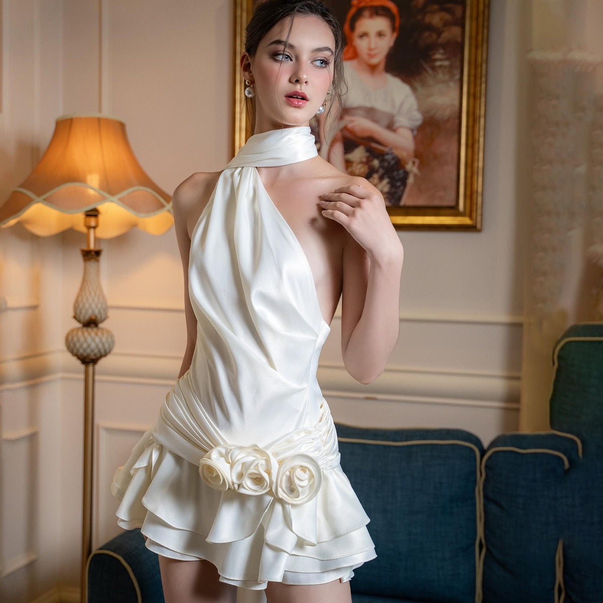 May include: A white satin halter dress with a draped bodice and a ruffled skirt. The dress features a floral embellishment at the waist. The model is wearing the dress in a room with a lamp and a painting.