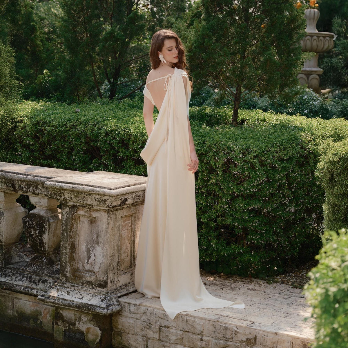 May include: A cream-colored, floor-length gown with a draped back and a flowing train. The dress features delicate straps and a soft, elegant silhouette. The woman is standing in a garden setting with lush greenery and a stone wall.
