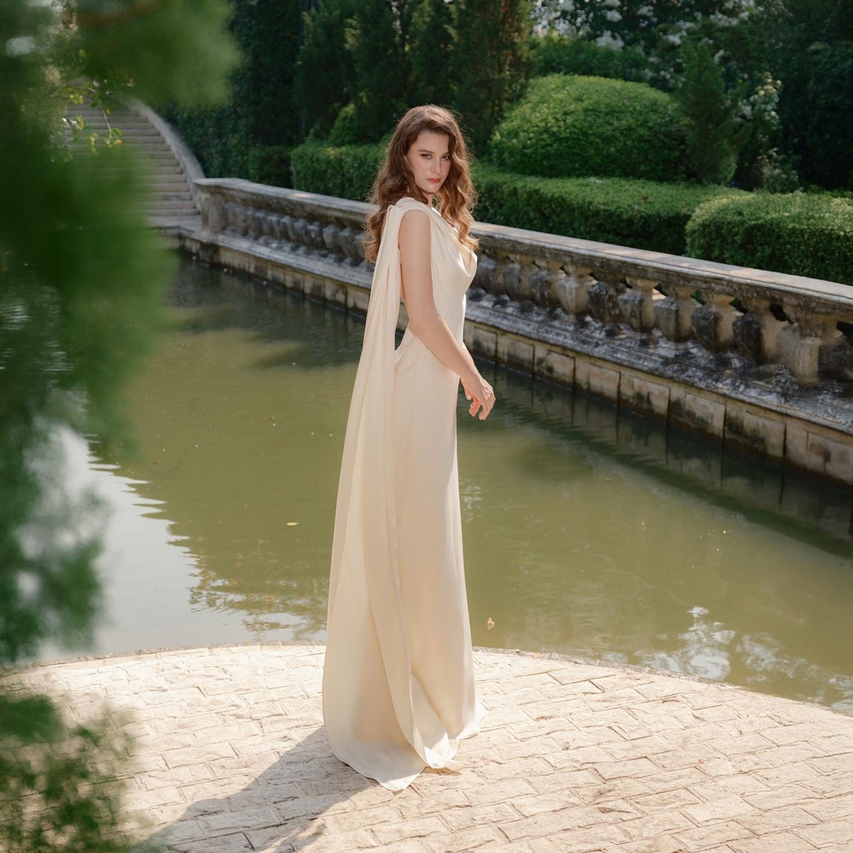 May include: A cream-colored, floor-length gown with a flowing cape detail. The dress has a simple, elegant design, perfect for a formal event. The woman is standing on a brick surface near a body of water, with greenery and a stone bridge in the background.