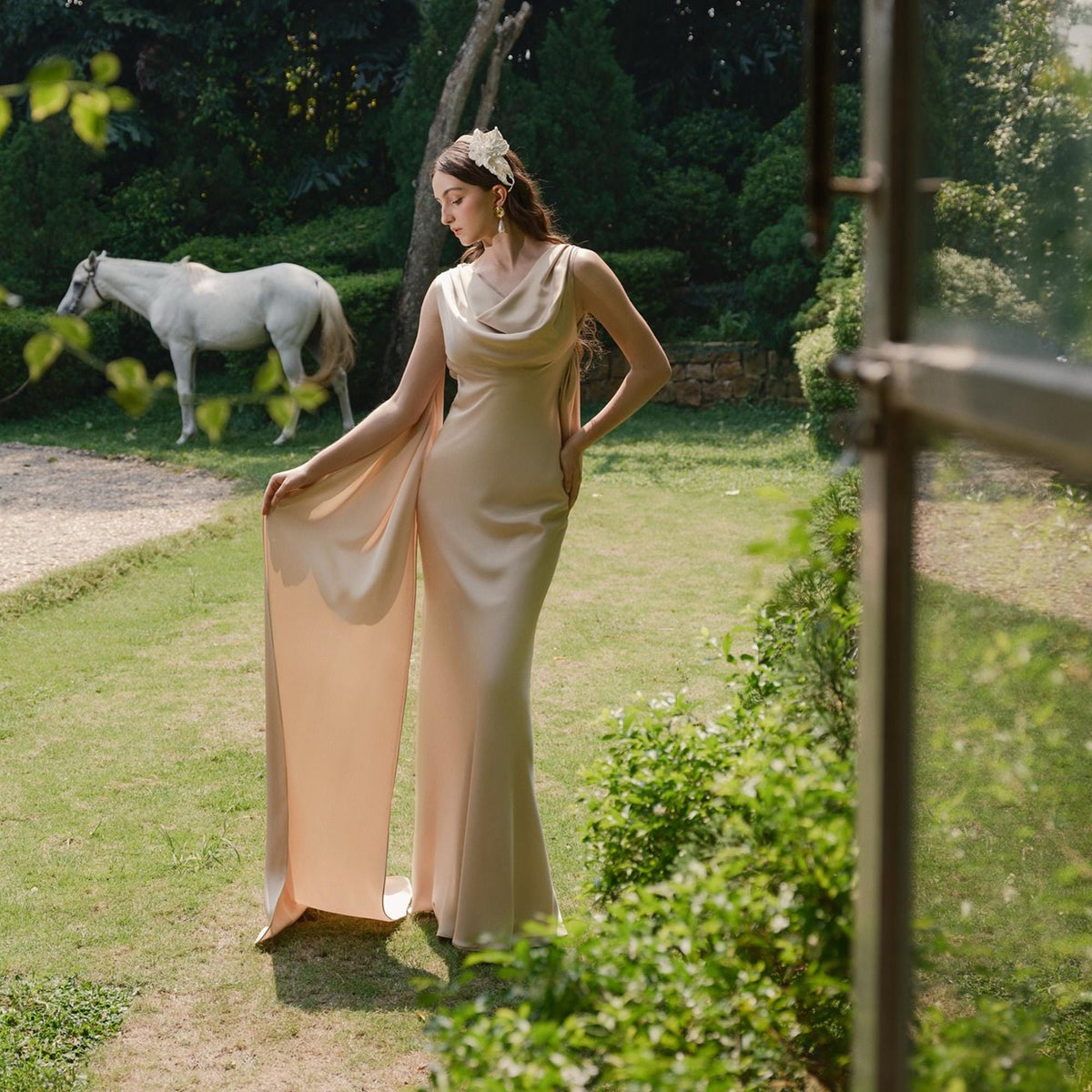 May include: A beige, floor-length gown with a cowl neckline and flowing cape sleeves. The dress is worn outdoors in a garden setting, with a white horse in the background. The dress is made of a silky material.