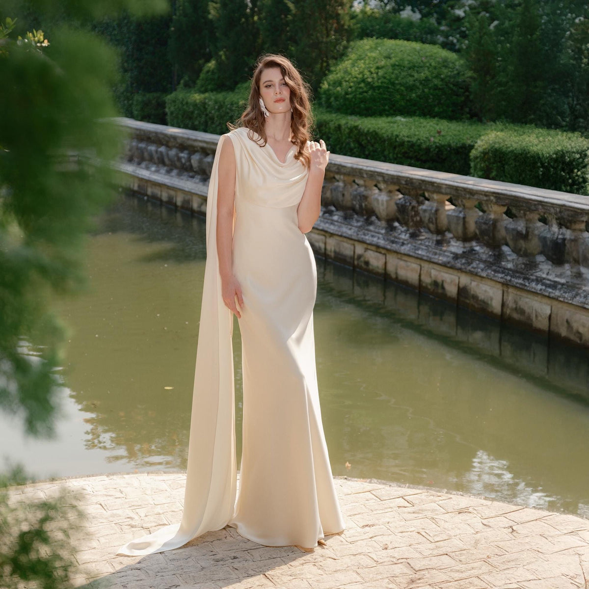 May include: A cream-colored, floor-length gown with a draped cowl neckline and a flowing cape detail. The dress is form-fitting through the bodice and flares out slightly at the hem. The image is set outdoors with a water feature and greenery in the background.