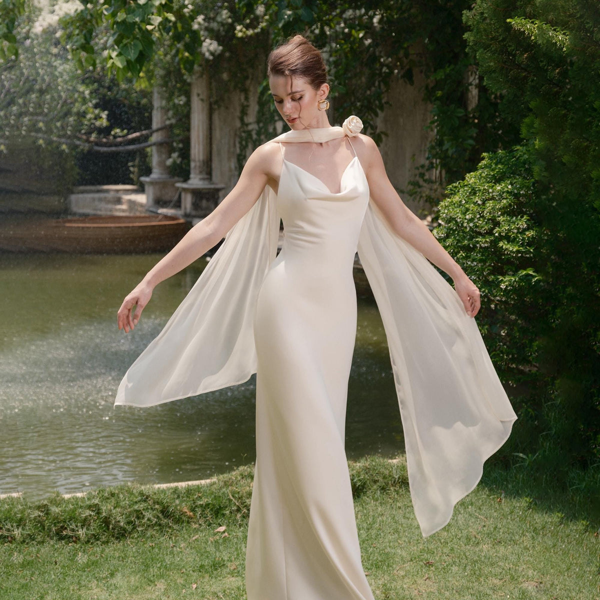 May include: A cream-colored, floor-length gown with a cowl neckline and spaghetti straps. The dress features sheer, flowing fabric attached at the shoulders, creating a cape-like effect. A small floral accent adorns the shoulder. The background includes a water feature and greenery.