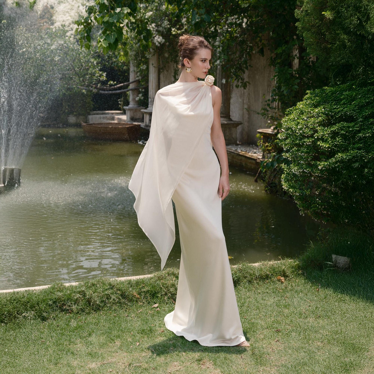 May include: A cream-colored, floor-length gown with a draped, asymmetrical cape. The dress features a high neckline and a floral accent on the shoulder. The background includes a fountain, greenery, and a pond, creating an elegant outdoor setting.