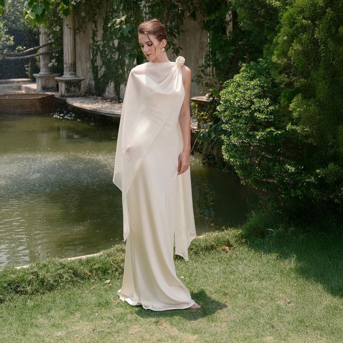 May include: A cream-colored, floor-length gown with a draped neckline and a sheer shawl. A fabric flower adorns the shoulder. The dress is worn outdoors near a pond and greenery.