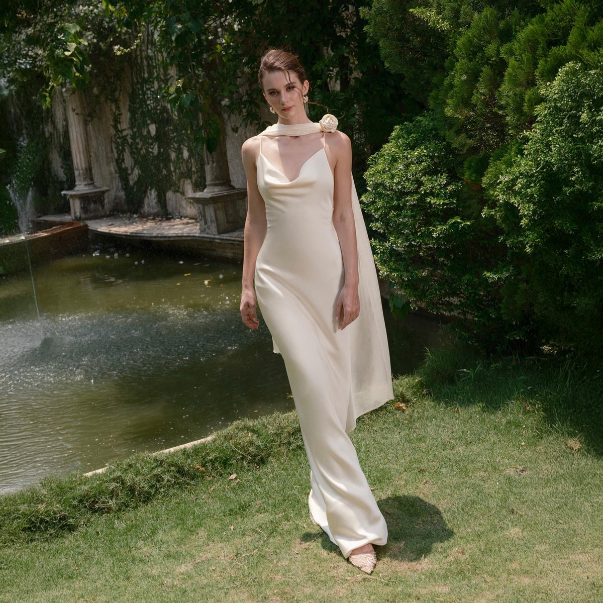 May include: A cream-colored, floor-length dress with spaghetti straps and a cowl neckline. A matching scarf is draped over the shoulders, secured with a floral accent. The dress is worn outdoors near a pond and lush greenery.