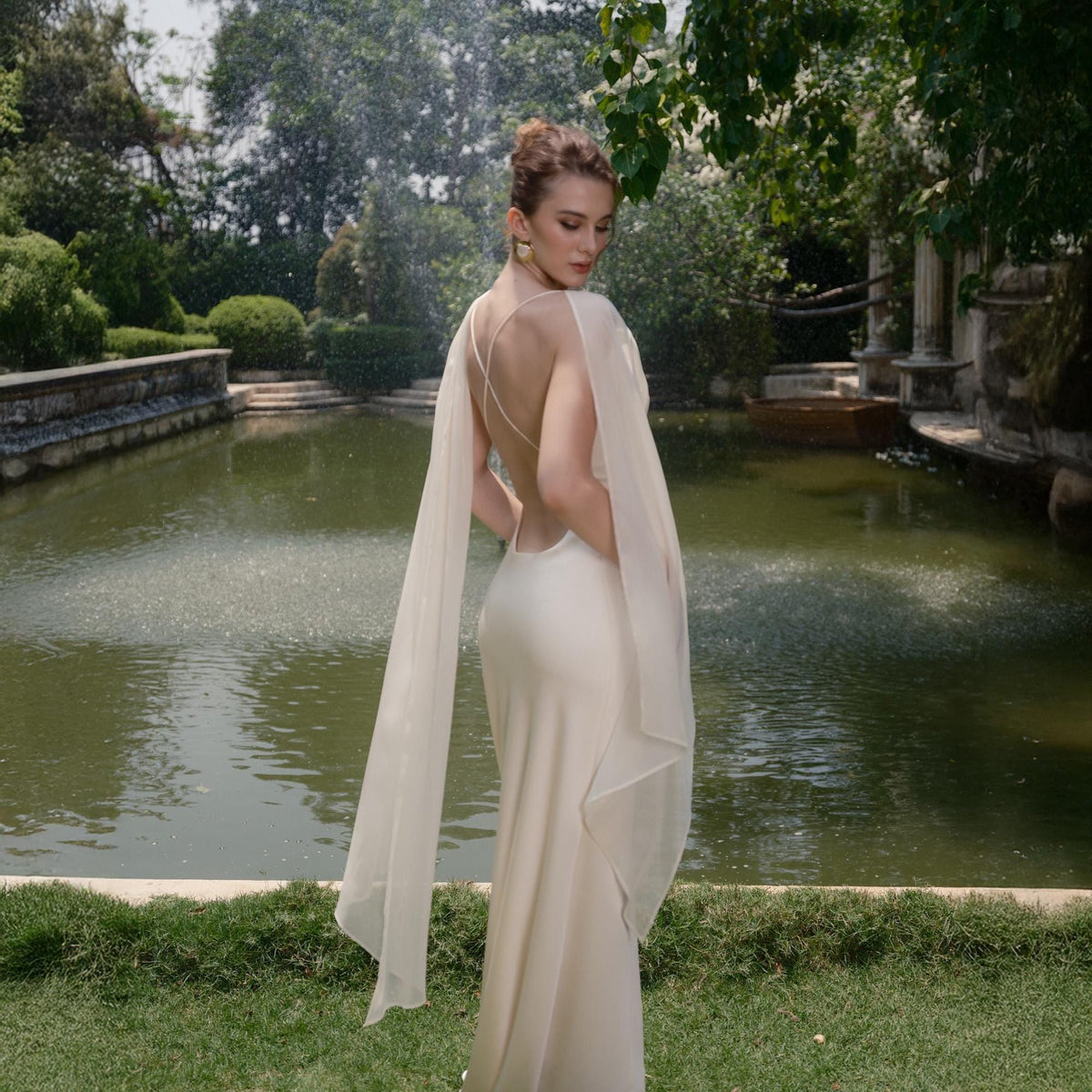 May include: A cream-colored, backless gown with delicate spaghetti straps and flowing chiffon sleeves. The dress is worn outdoors near a pond with a fountain. The model is wearing gold earrings and has her hair up.