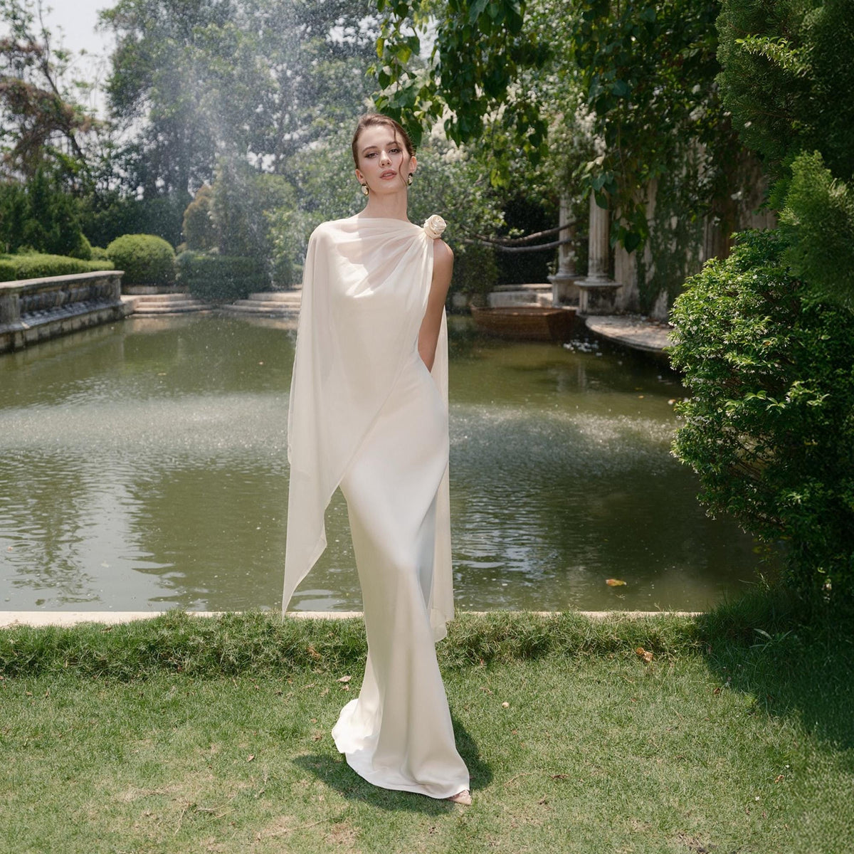 May include: A floor-length ivory gown with a draped, off-the-shoulder design. The dress features a flowing cape-like overlay and a small floral accent at the shoulder. The model is standing in front of a pond with a fountain, surrounded by greenery.