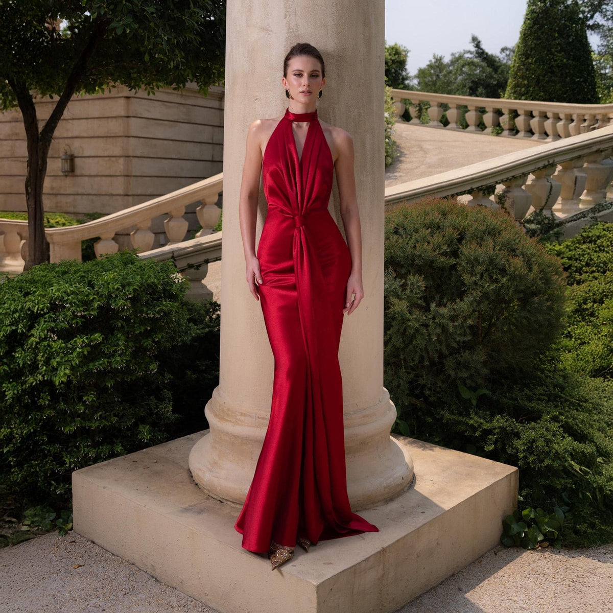 May include: A floor-length, red satin gown with a halter neckline and a knot detail at the waist. The dress has a flowing skirt and is worn in an outdoor setting. The model is standing next to a stone pillar.