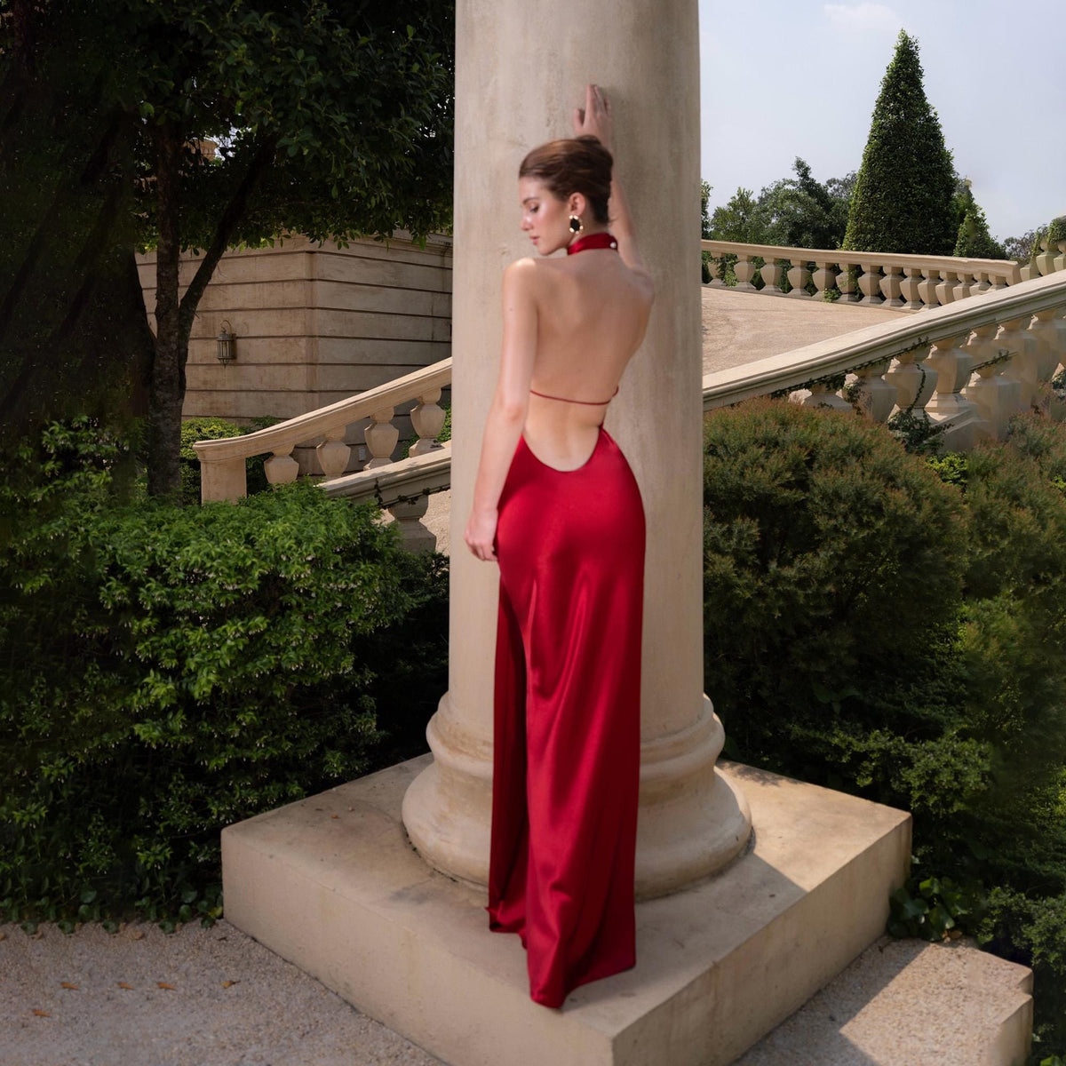 May include: A vibrant red, backless evening gown with a halter neckline and a high slit. The dress is made of a silky material and drapes elegantly. The model is standing next to a large, white column in an outdoor setting.