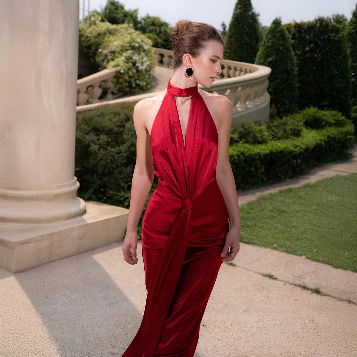 May include: A vibrant red halter dress with a flowing design. The dress features a high neckline and a deep V-neck, with a knot detail at the waist. The dress is made of a shiny, silky material. Black earrings complete the look.