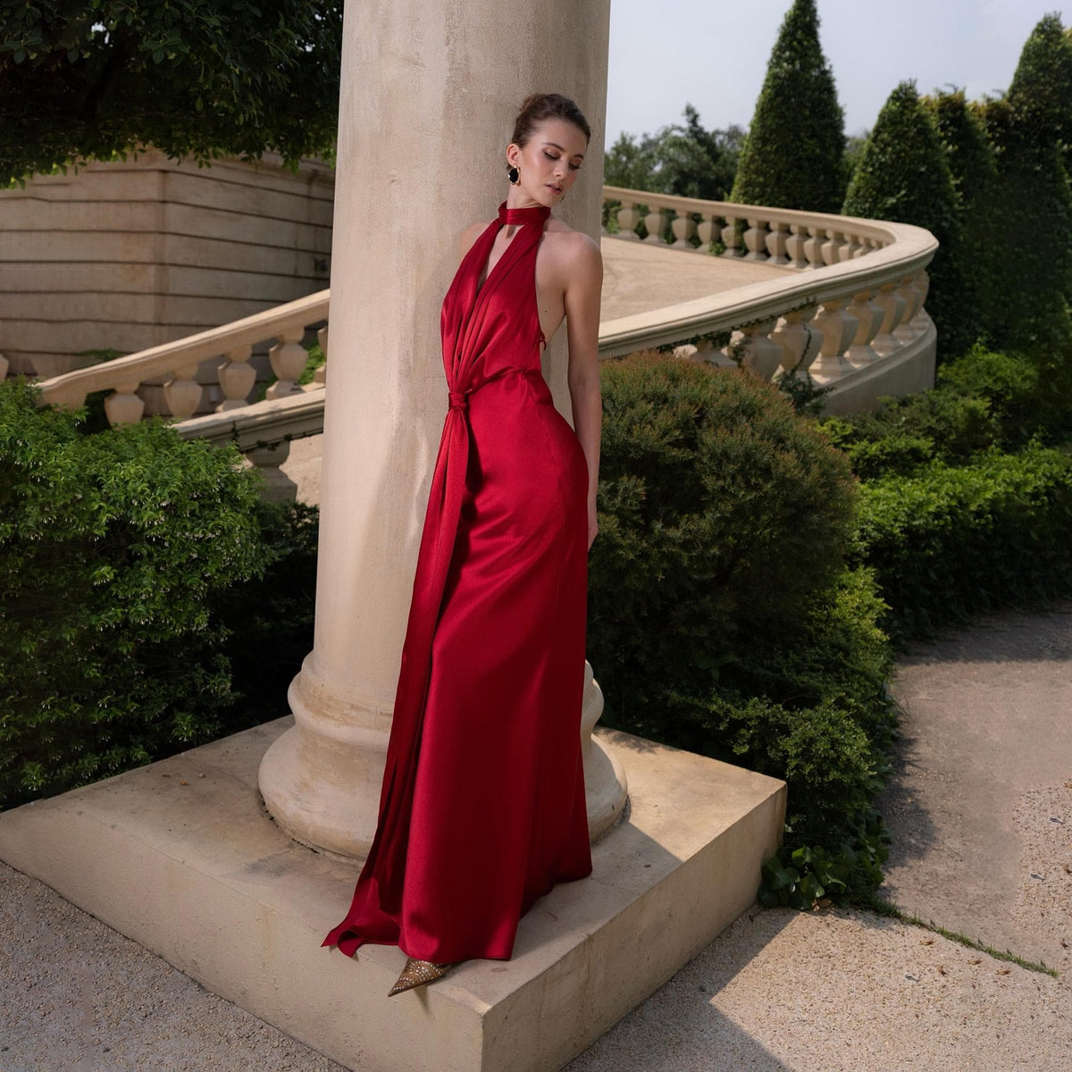 May include: A long, flowing red satin gown with a halter neckline and a draped design. The dress features a floor-length skirt and is worn outdoors near a stone pillar and a garden.