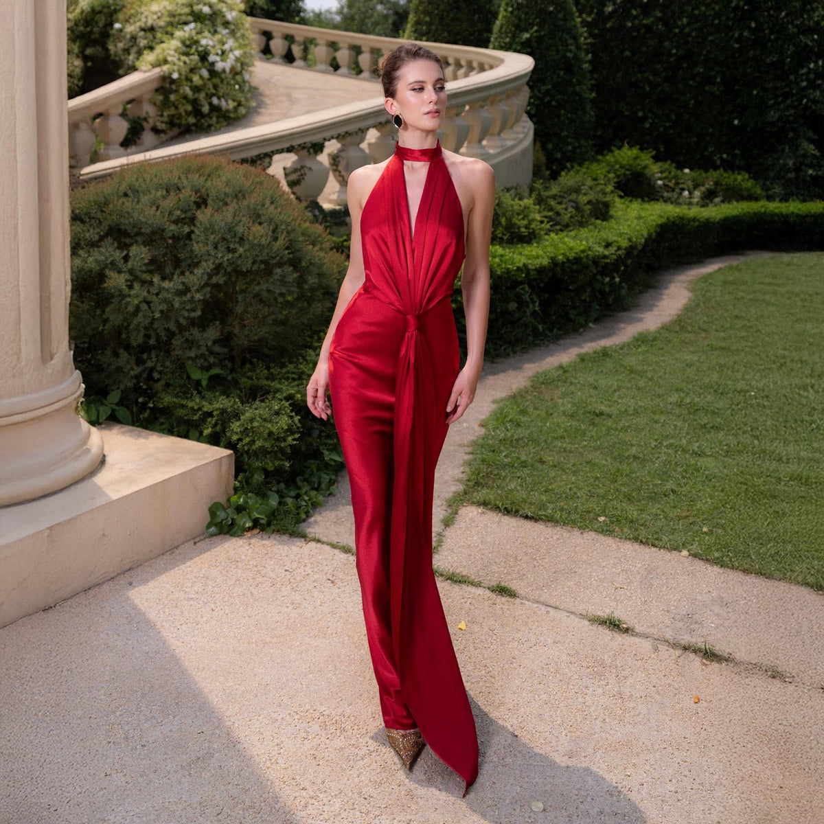 May include: A red halter jumpsuit with a flowing, wide-leg design. The jumpsuit features a high neckline and a knot detail at the waist. The model is standing on a stone path in a garden setting.