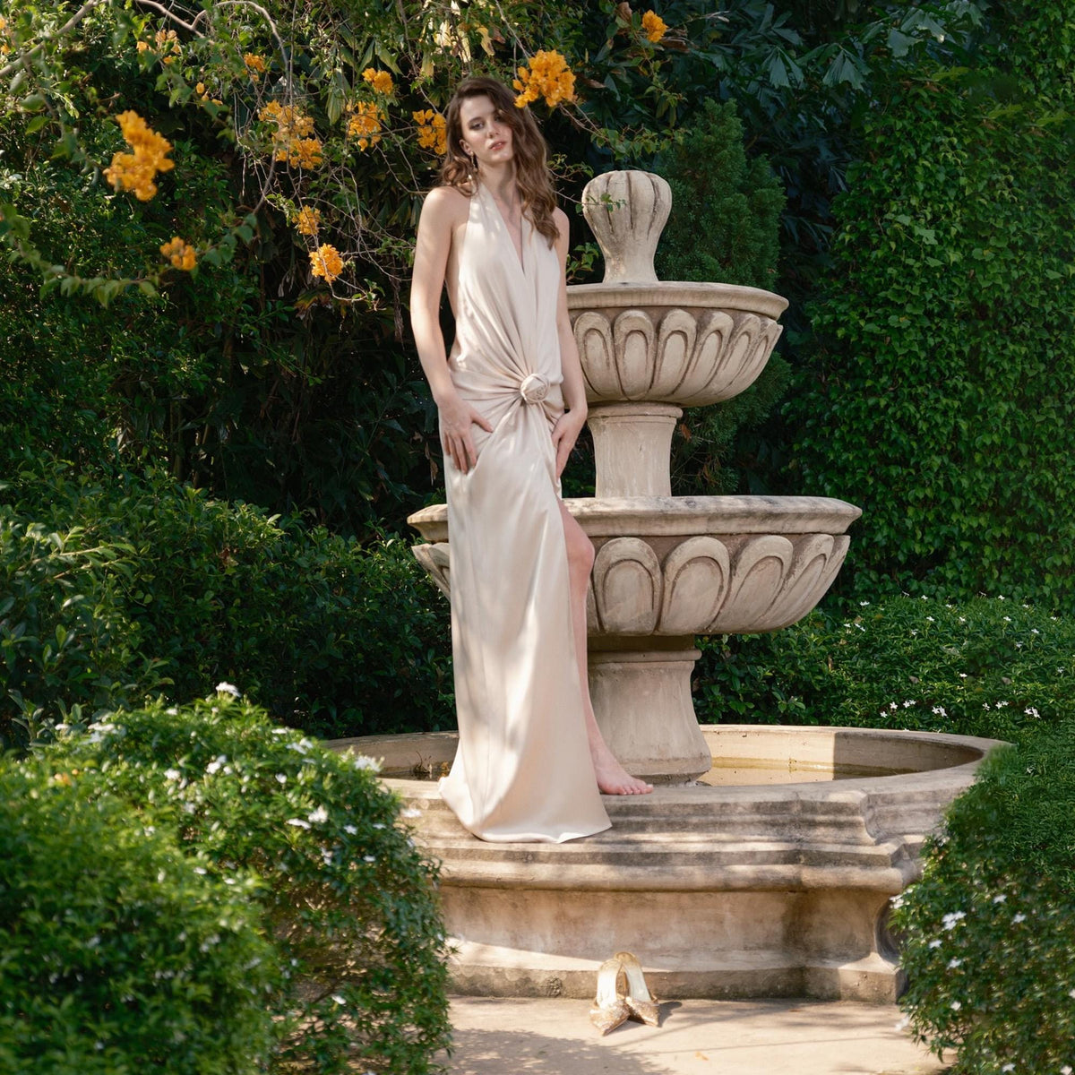 May include: A beige halter-neck gown with a knot detail at the waist and a thigh-high slit. The dress is worn outdoors near a stone fountain and lush greenery. The gown's fabric drapes elegantly, creating a flowing silhouette.