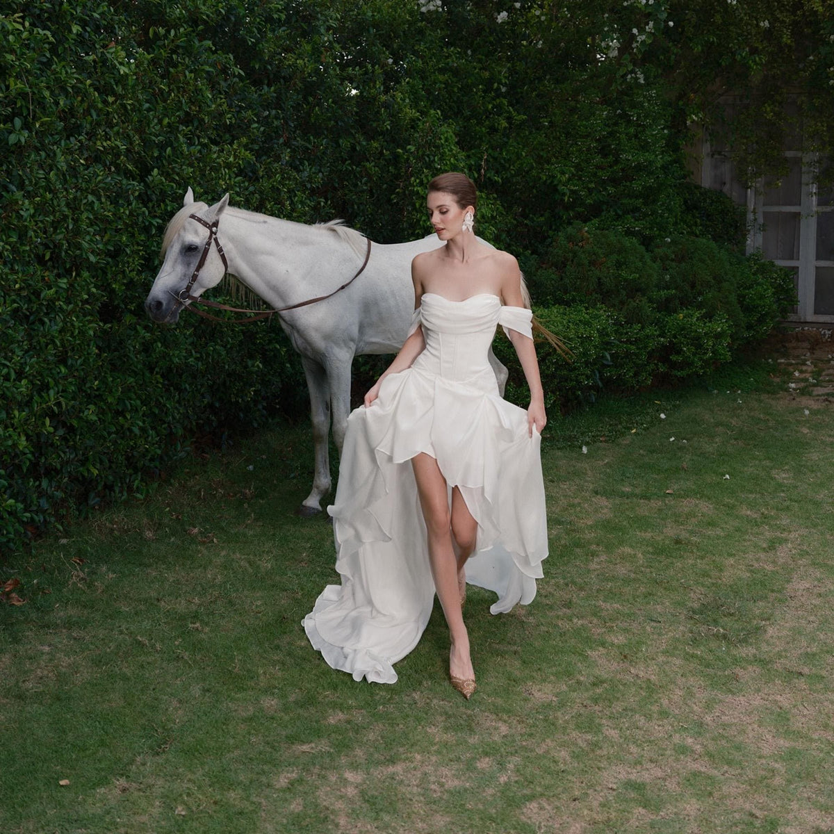 May include: A white, off-the-shoulder wedding dress with a high-low hemline. The dress features a fitted bodice and flowing skirt. A white horse stands in the background near a green hedge. The scene is set outdoors on a grassy lawn.
